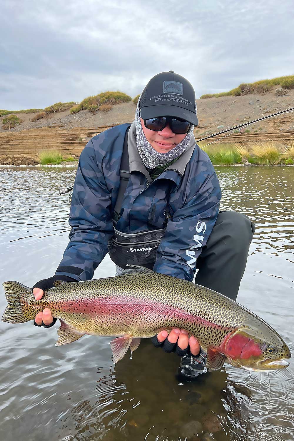 An angler in outdoor gear kneels in shallow water, proudly holding a large rainbow trout with vibrant red and green coloring, with grassy banks and overcast sky in the background. | Belize River Lodge Guided Fishing Trips | Wildside Adventures Travel