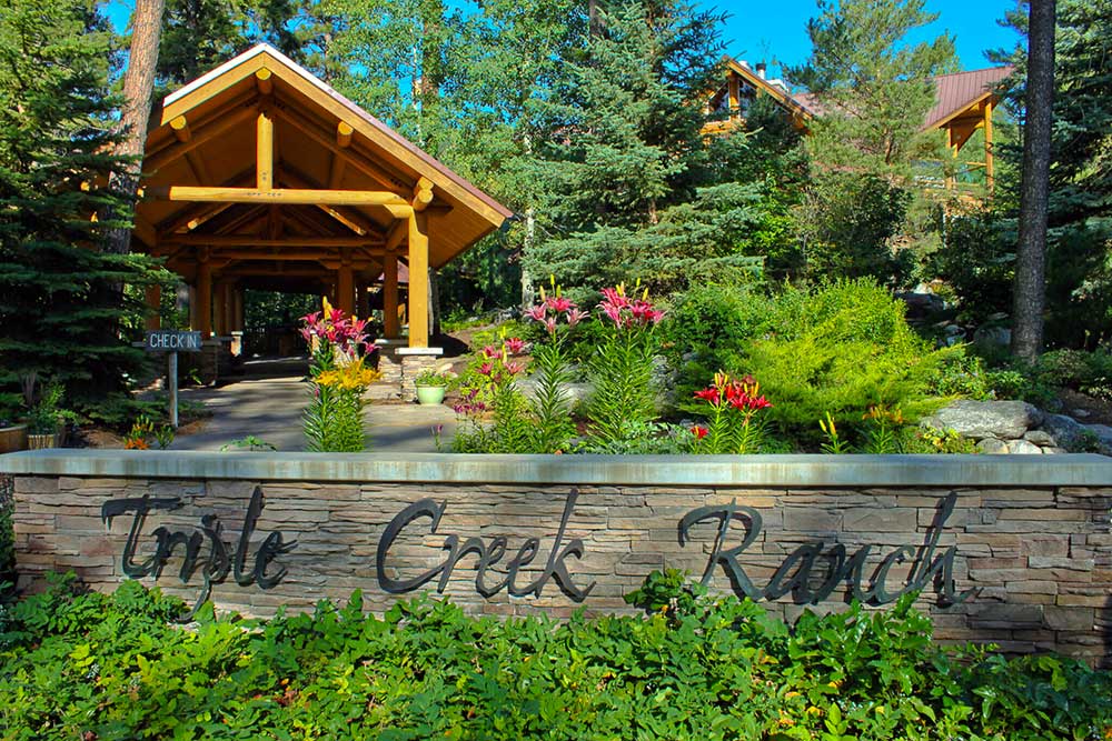 Entrance to Triple Creek Ranch with a stone sign amidst lush greenery and blooming flowers