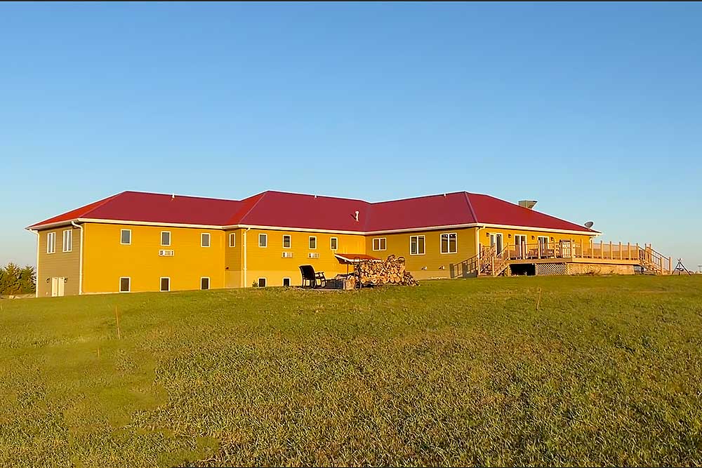 A large yellow building with a red roof sits on a grassy hill under a clear blue sky, serving as a welcoming South Dakota bird lodge. An outdoor seating area and stacked firewood add charm to this scenic spot. | United States Bird Hunting Trips| Wildside Adventures Travel