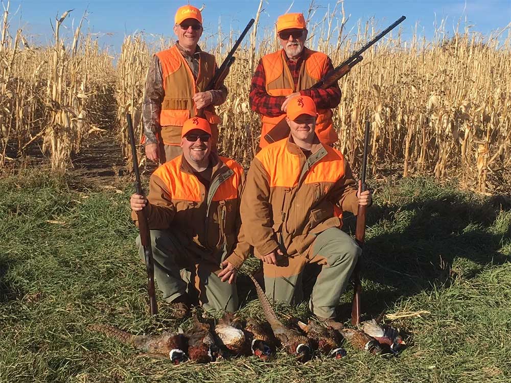 dakota-prairie-lodge-13 Four hunters in orange vests and hats pose by a cornfield, holding shotguns. Several pheasants are laid out on the grass—another successful day at Dakota Prairie Lodge hunting, a premier South Dakota bird lodge experience. | United States Bird Hunting Trips| Wildside Adventures Travel