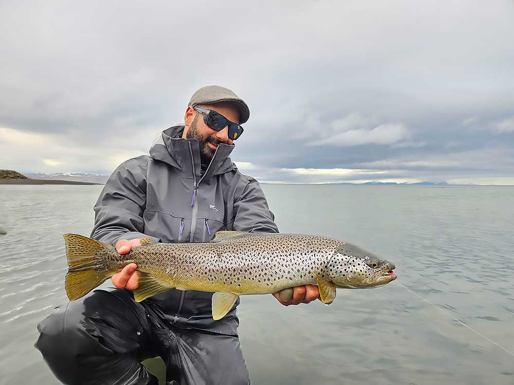 long-valley-lodge-image-2 Joe Rossi grins as he stands in shallow water, holding a large spotted fish after a successful Wildside Adventures Iceland salmon fishing trip. He sports sunglasses, a cap, and a gray jacket with mountains and clouds in the background. | Belize River Lodge Guided Fishing Trips | Wildside Adventures Travel