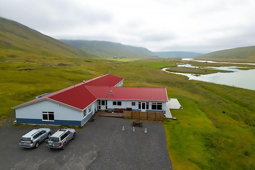 long-valley-lodge-top-2 A building with a red roof and white walls sits in a grassy valley near a river, with parked cars in front and green hills under a cloudy sky in the background. | Belize River Lodge Guided Fishing Trips | Wildside Adventures Travel