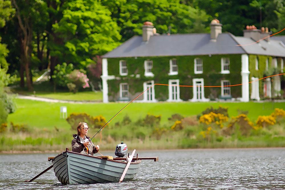 A person sits in a small rowboat on a lake, fly fishing. Lush greenery and a large ivy-covered house with chimneys are visible in the background. The scene is peaceful and outdoorsy. | Belize River Lodge Guided Fishing Trips | Wildside Adventures Travel