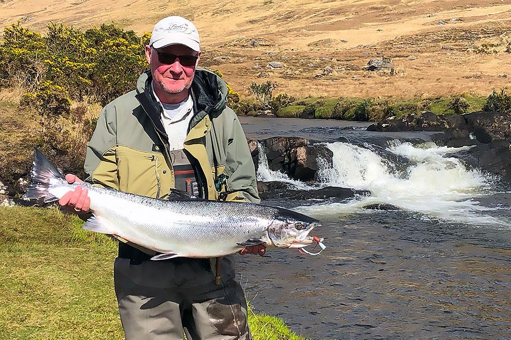 A man wearing sunglasses and outdoor fishing gear smiles while holding a large silver fish by a river, with a small waterfall and grassy, rocky landscape in the background. | Belize River Lodge Guided Fishing Trips | Wildside Adventures Travel