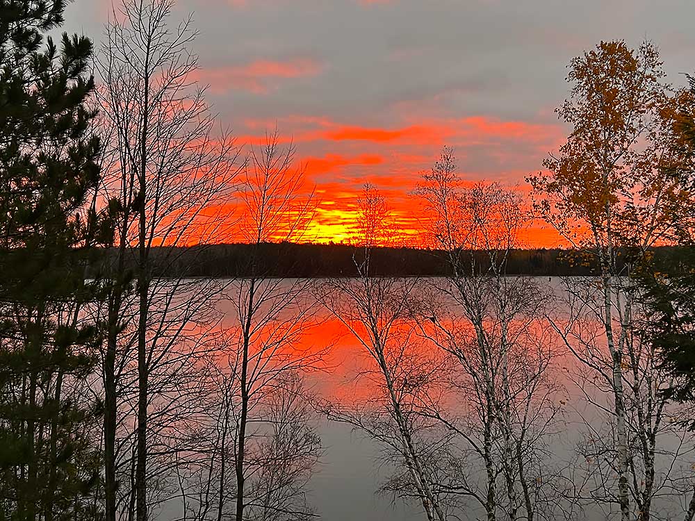 Flambeau-13 Bare trees stand by a calm lake reflecting a vivid orange sunset, with dark clouds overhead and a silhouetted forest on the horizon. | Belize River Lodge Guided Fishing Trips | Wildside Adventures Travel
