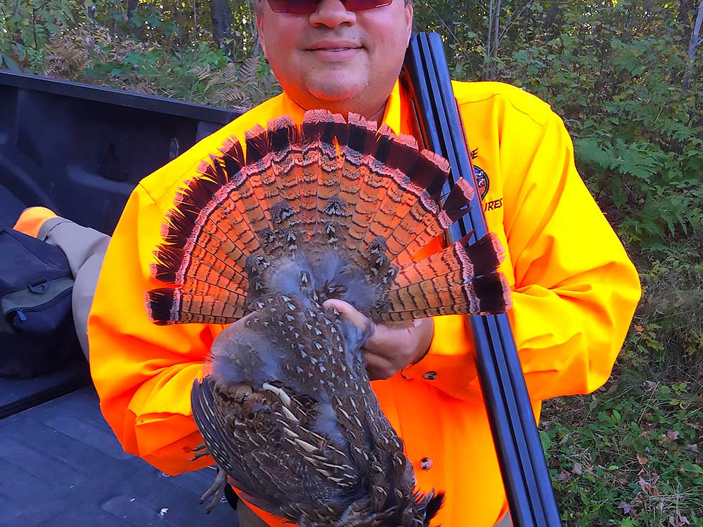 Flambeau-16 A person in a bright orange jacket and sunglasses holds up a ruffed grouse with its tail feathers fanned out. The person is standing beside a vehicle in a wooded area. | Belize River Lodge Guided Fishing Trips | Wildside Adventures Travel