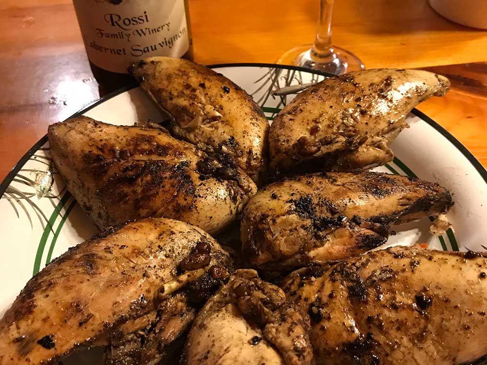 Flambeau-17 A plate of grilled, seasoned chicken breasts arranged in a circle, with a bottle of Rossi Family Winery Cabernet Sauvignon and a wine glass in the background on a wooden table. | Belize River Lodge Guided Fishing Trips | Wildside Adventures Travel