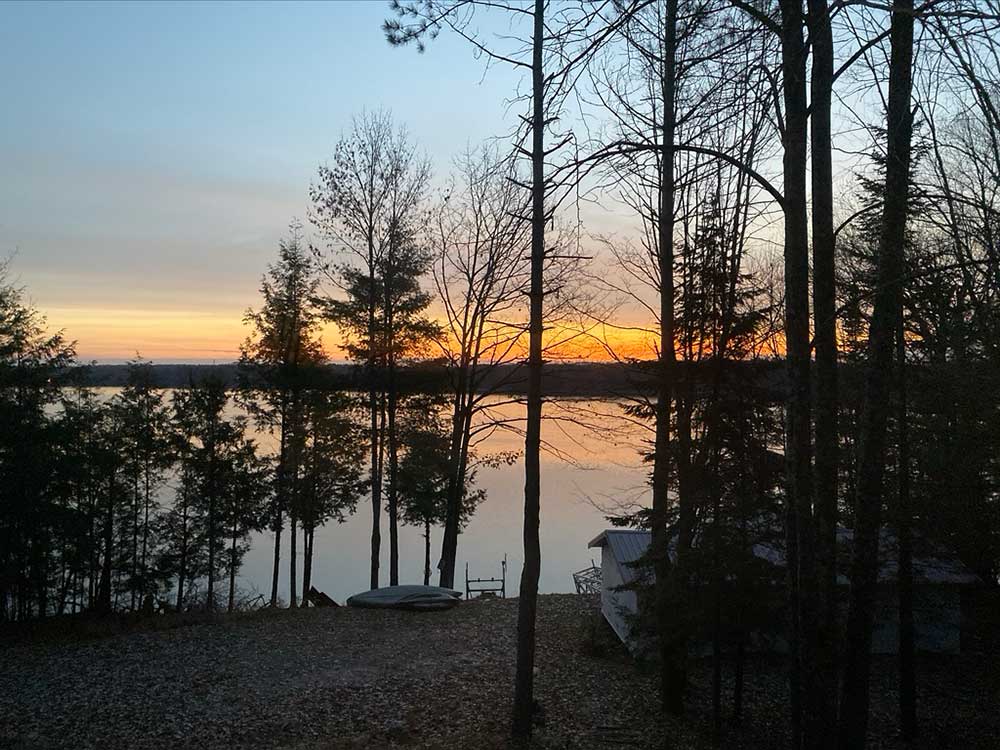 Flambeau-18 Sunset over a calm lake, with bare and evergreen trees in the foreground. A small dock and boat are visible near the water, and a cabin sits to the right among the trees. The sky glows orange and blue. | Belize River Lodge Guided Fishing Trips | Wildside Adventures Travel