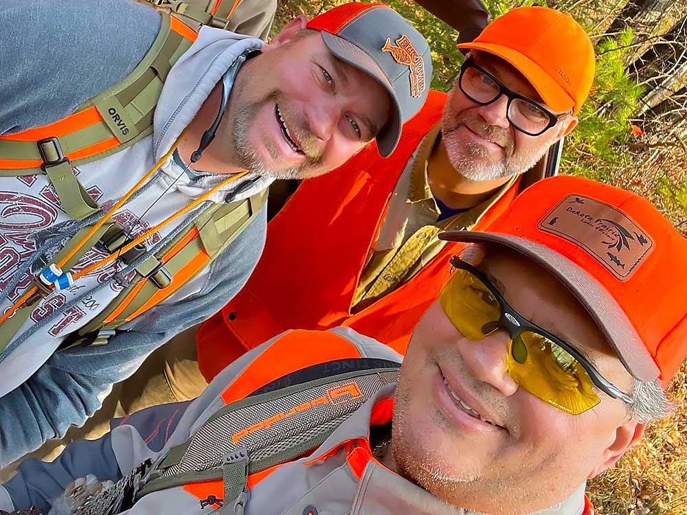 Flambeau-21 Three smiling men outdoors in hunting gear, including orange vests, hats, and sunglasses. They appear to be on a hiking or hunting trip, surrounded by trees and natural light. | Belize River Lodge Guided Fishing Trips | Wildside Adventures Travel
