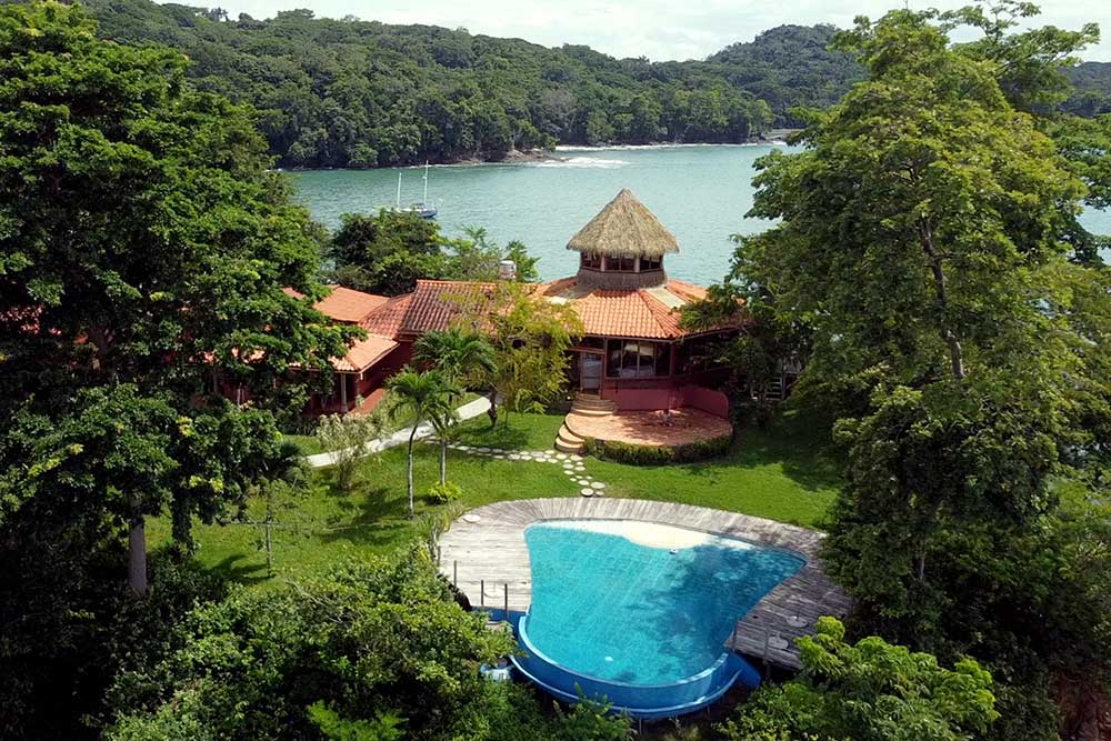A tropical villa with a thatched roof sits beside a turquoise pool, surrounded by lush greenery and trees, overlooking a calm bay with forested hills in the background. | Belize River Lodge Guided Fishing Trips | Wildside Adventures Travel