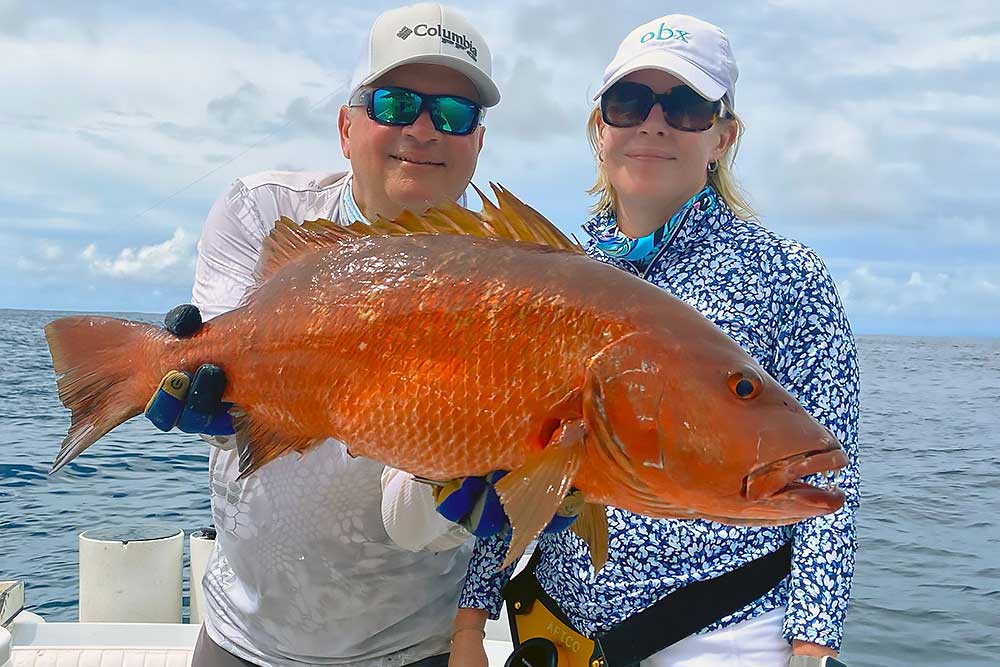 Two people on a boat hold up a large, reddish-orange fish they caught. Both are wearing sunglasses, hats, and patterned, sun-protective clothing. The ocean and cloudy sky are visible in the background. | Belize River Lodge Guided Fishing Trips | Wildside Adventures Travel