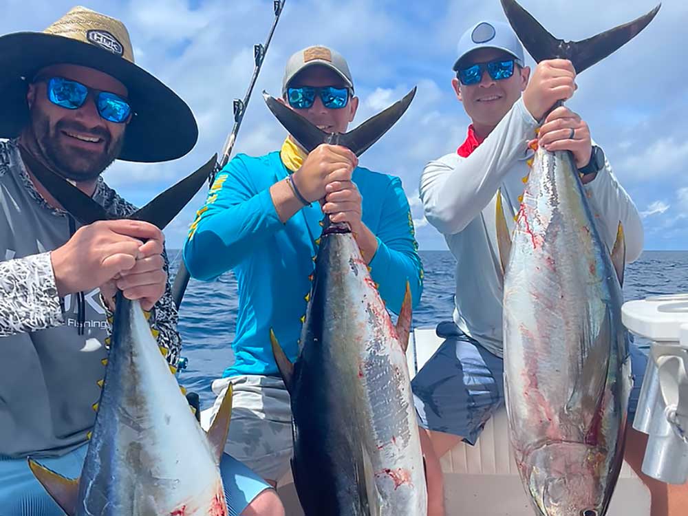 Three men on a boat proudly display large tuna catches under a blue sky, epitomizing the thrilling fishing trips offered by Wildside Adventures in stunning outdoor destinations.| Wildside Adventures | Fishing and Hunting Trips