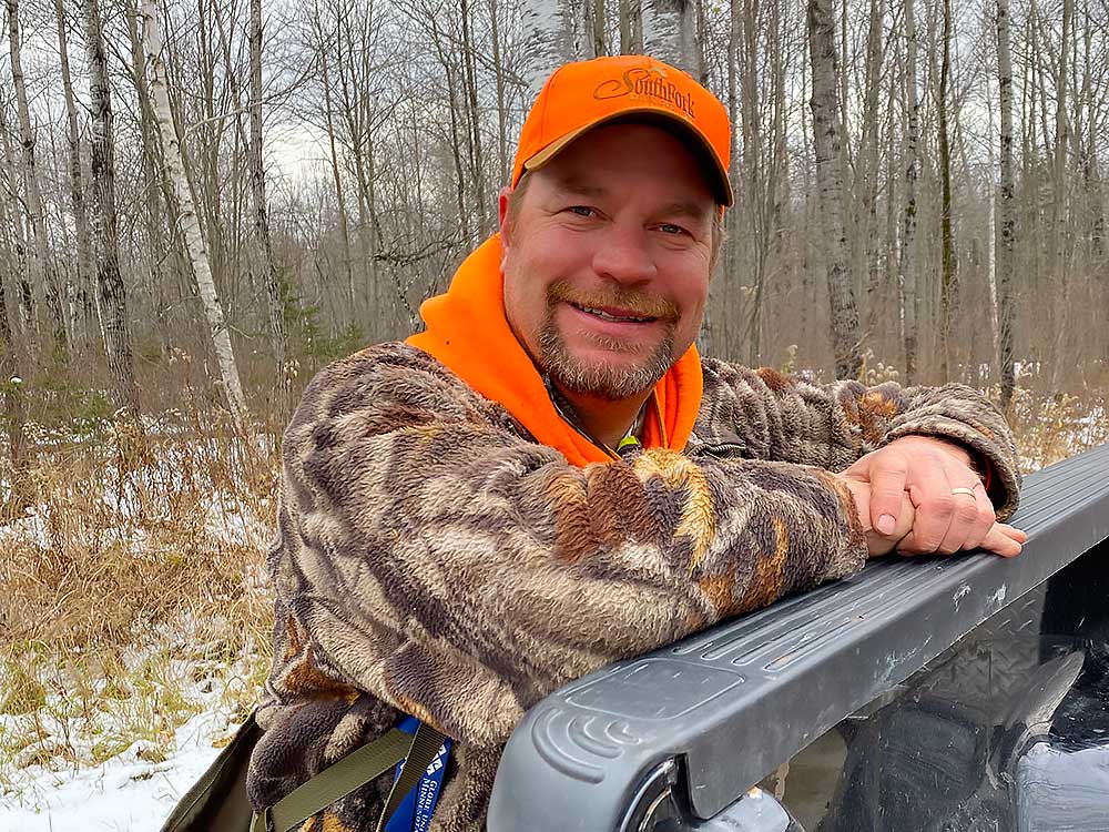flambeau-top-1b A man wearing an orange cap and camouflage jacket smiles while leaning on the side of a black vehicle in a snowy, leafless forest. | Belize River Lodge Guided Fishing Trips | Wildside Adventures Travel